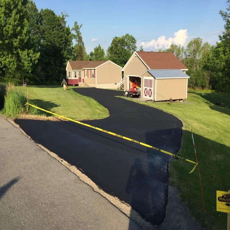 Freshly sealcoated curving driveway blocked off with caution tape while it cures, with the house and garage behind it